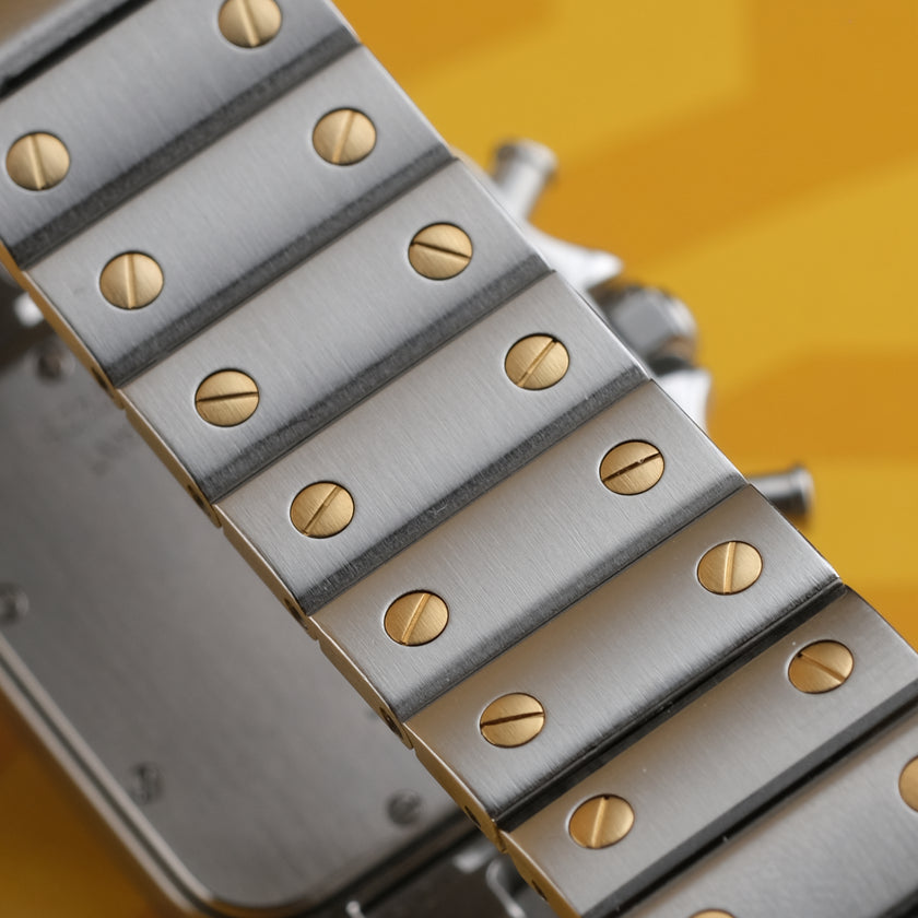 A Cartier watch with a silver-toned stainless steel case and bracelet, a gold-toned bezel, and a white dial with Roman numerals, set against a yellow background.