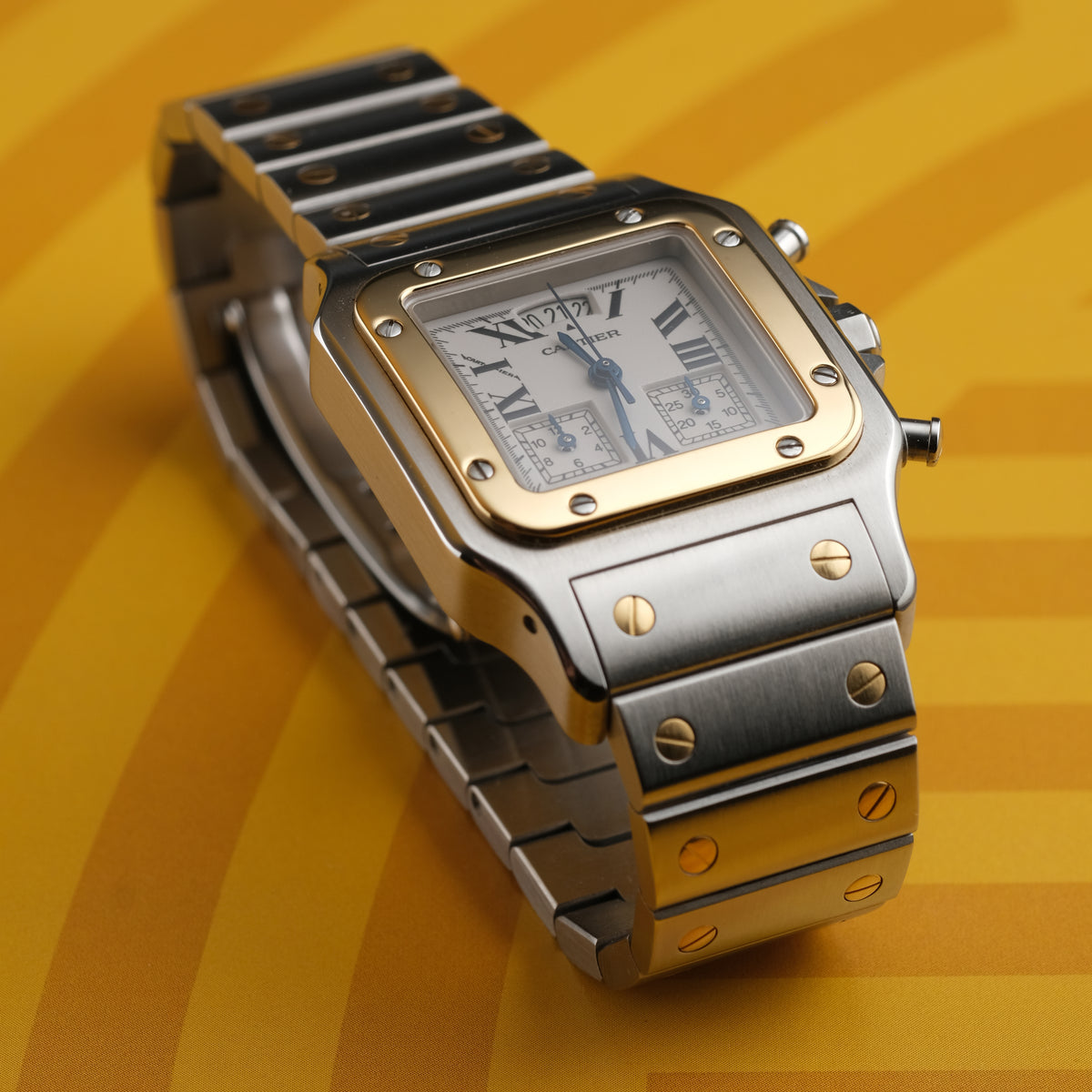 A Cartier watch with a silver-toned stainless steel case and bracelet, a gold-toned bezel, and a white dial with Roman numerals, set against a yellow background.