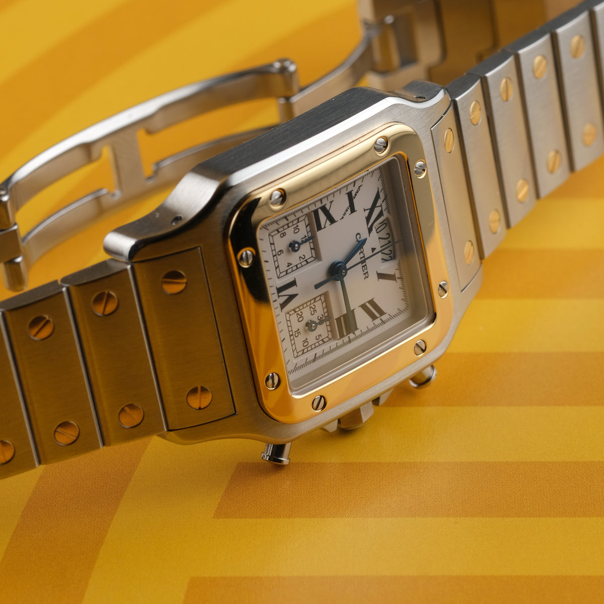 A Cartier watch with a silver-toned stainless steel case and bracelet, a gold-toned bezel, and a white dial with Roman numerals, set against a yellow background.