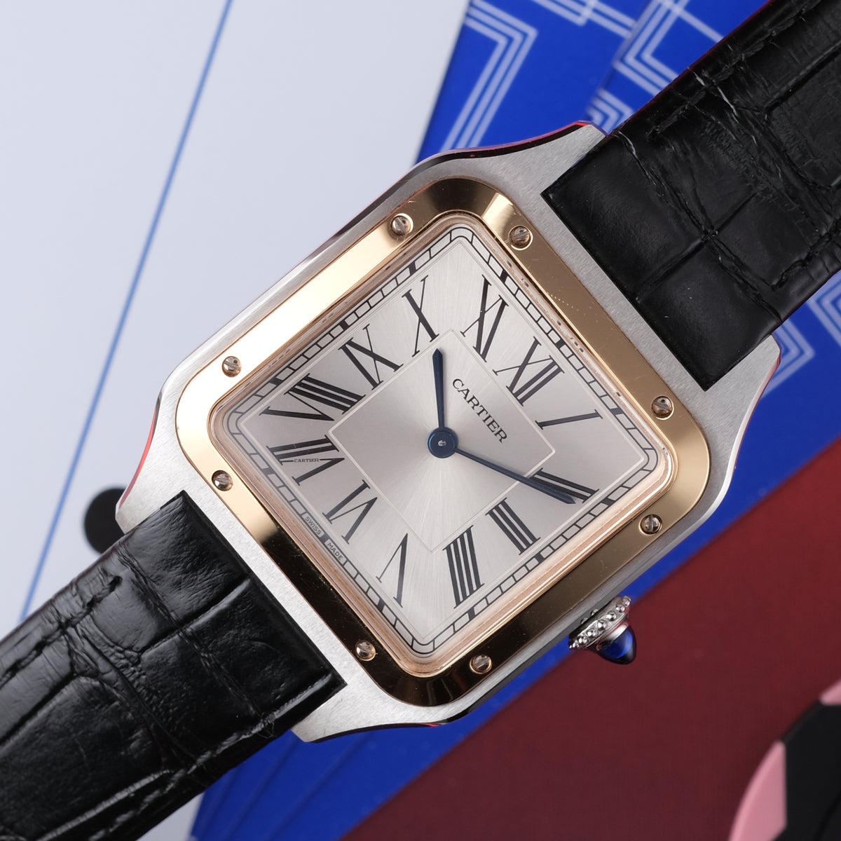 A Cartier watch with a square silver-toned case, gold-tone bezel, white dial with black Roman numerals, and a black crocodile-embossed leather strap, shown close-up against a graphic blue, red, and white background.