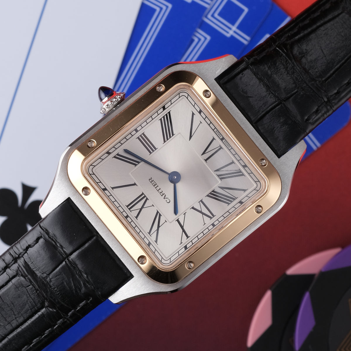 A Cartier Santos watch with a silver-toned stainless steel case, gold-tone bezel, white dial with Roman numerals, and a black alligator leather strap, shown against a stylized playing card background.