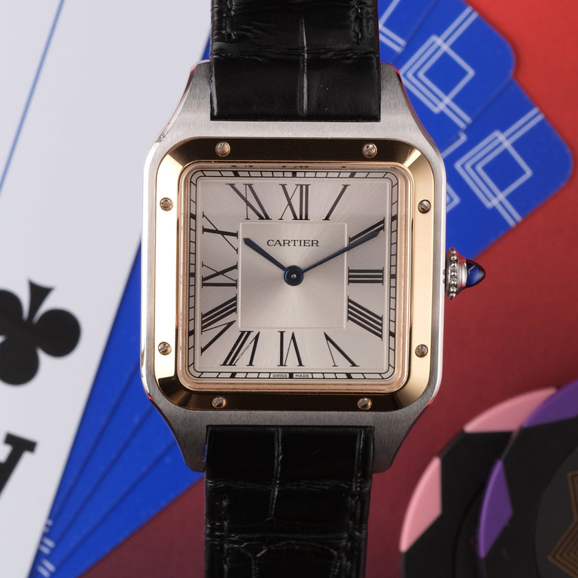 A Cartier Santos watch with a square stainless steel case, gold-tone bezel accents, a white dial with black Roman numerals, and a black alligator leather strap. The watch is centered against a colorful background featuring playing card suits.