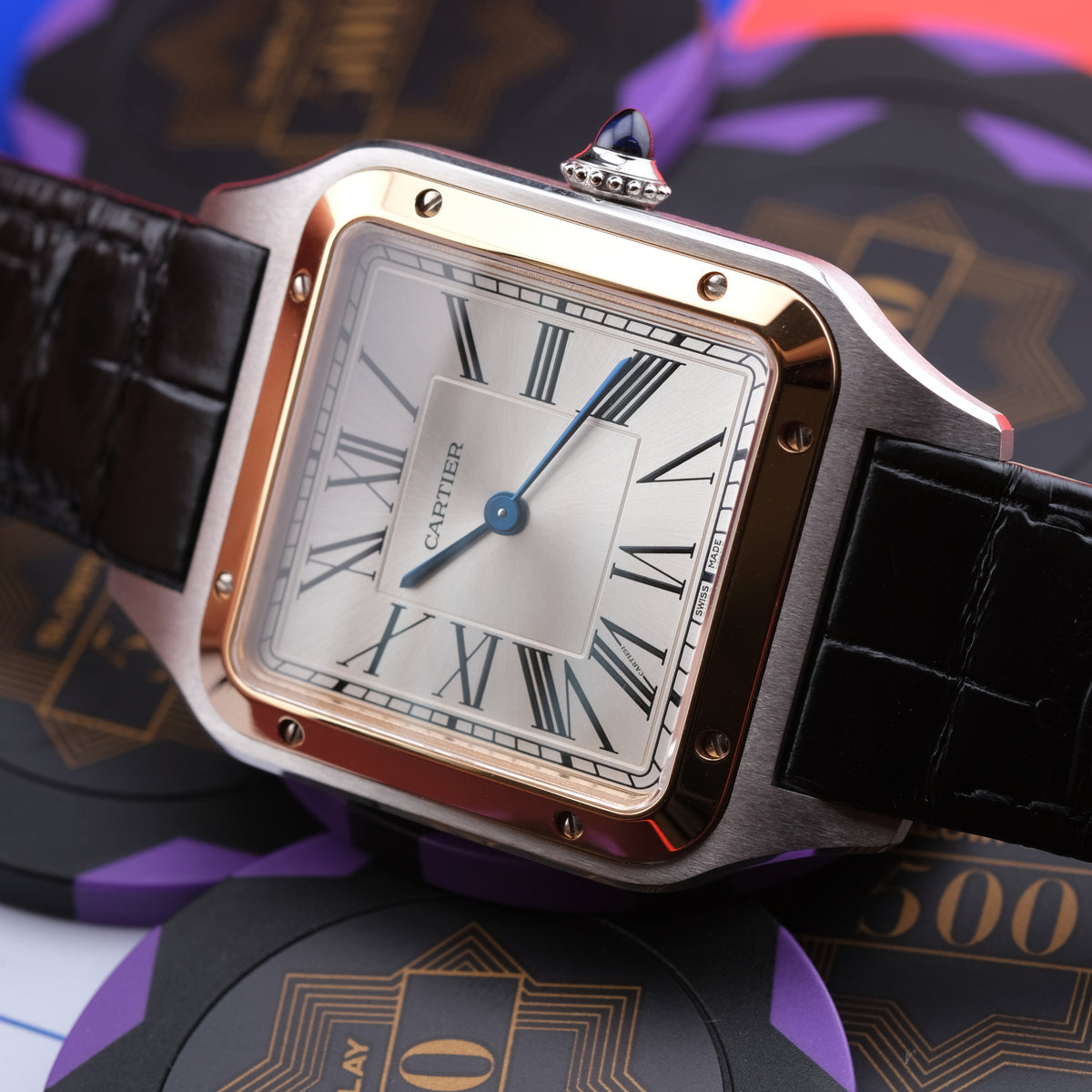 A Cartier Santos watch with a square silver-toned case, rose gold accents, a white dial with black Roman numerals, and a black leather strap, styled on top of purple and black branded cards.