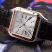 A Cartier Santos watch with a square silver-toned case, rose gold accents, a white dial with black Roman numerals, and a black leather strap, styled on top of purple and black branded cards.
