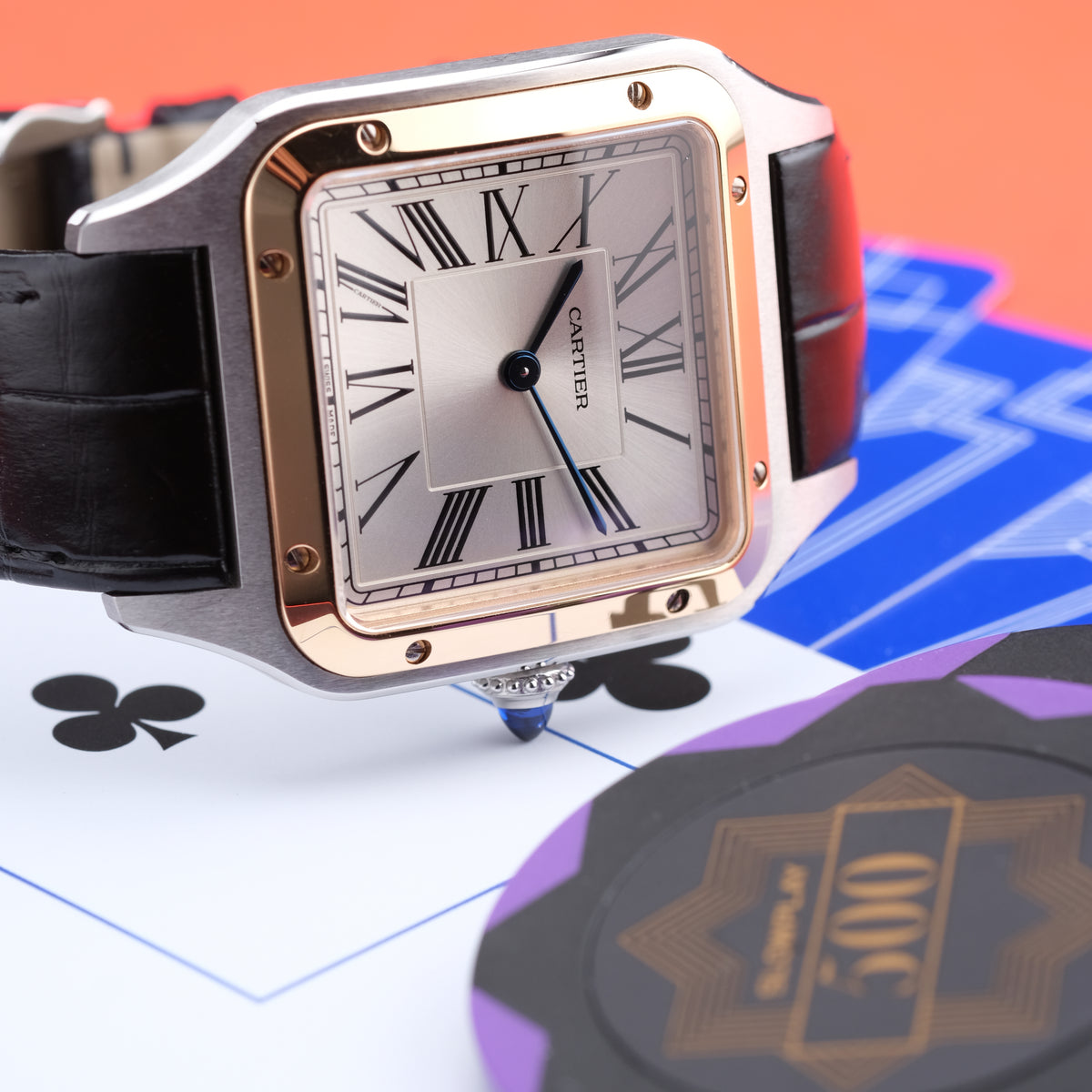 A Cartier watch with a square silver dial, rose gold-toned case, and black leather strap, styled on playing cards with poker chips in the foreground against an orange background.