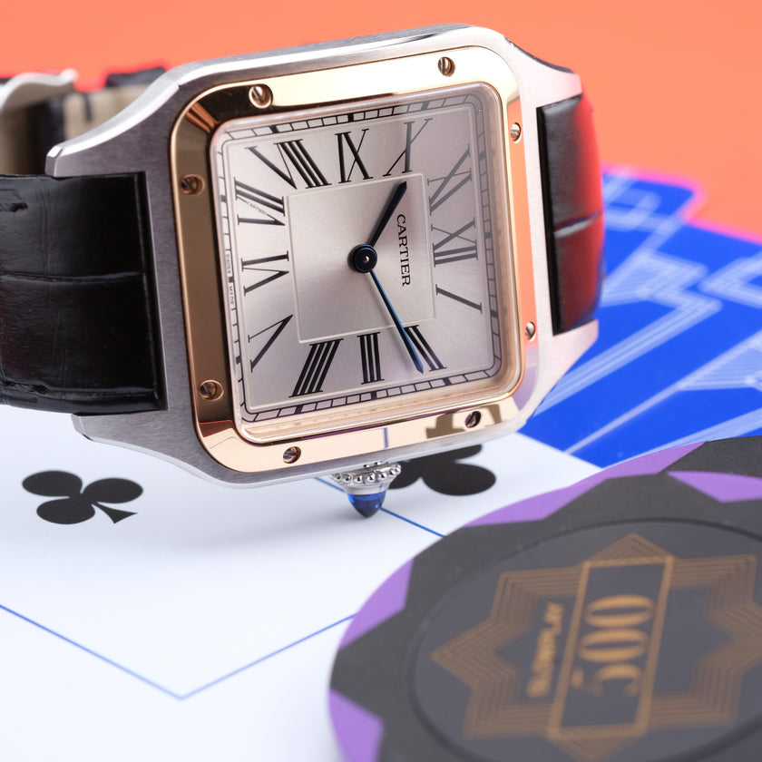 A Cartier watch with a square silver dial, rose gold-toned case, and black leather strap, styled on playing cards with poker chips in the foreground against an orange background.