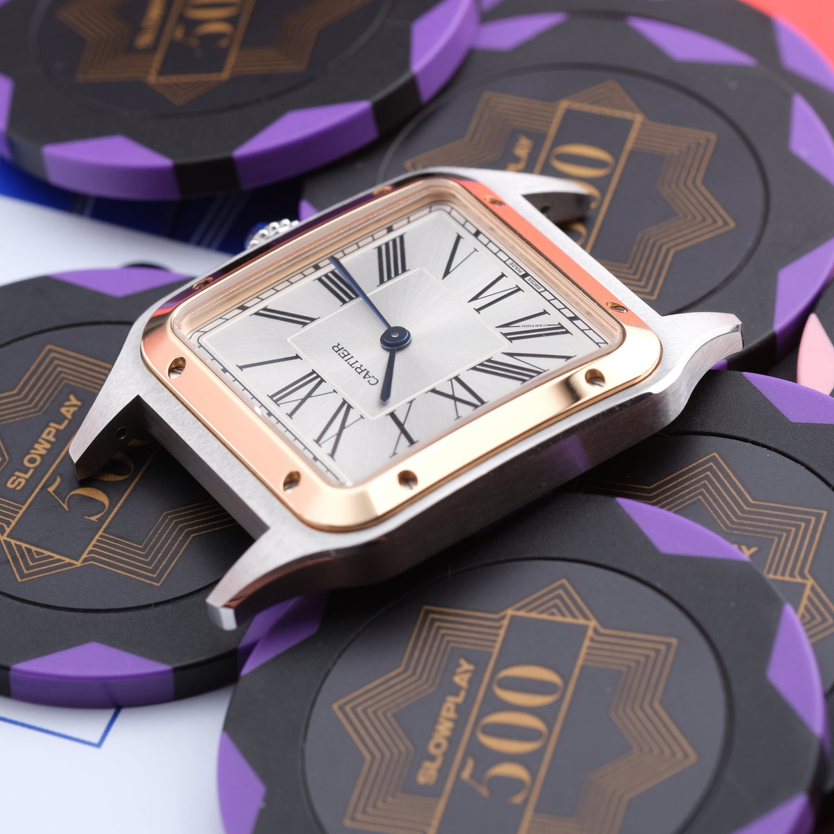 A rectangular analog watch with a silver-toned case, rose gold-toned bezel, and white dial with Roman numerals, styled on top of black and purple poker chips.