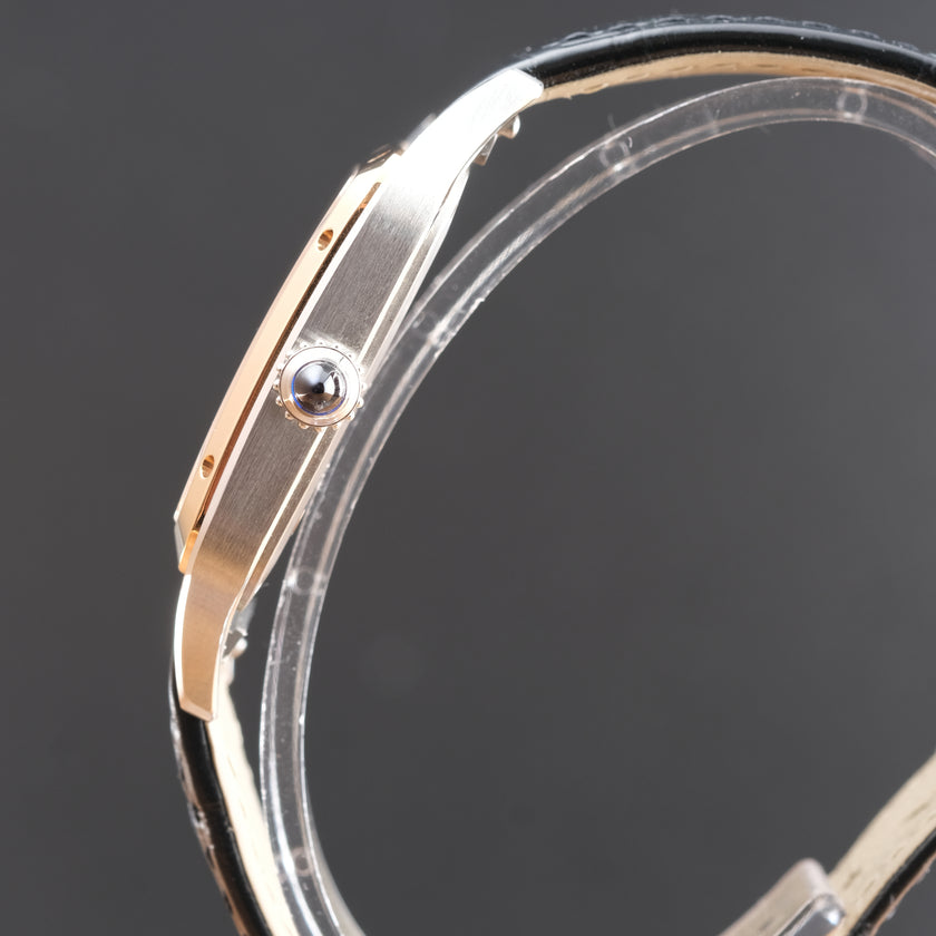 Close-up side view of a wristwatch with a silver-toned metal case, a rose gold-toned bezel accent, and a black leather strap, mounted on a clear display stand against a dark gray background.