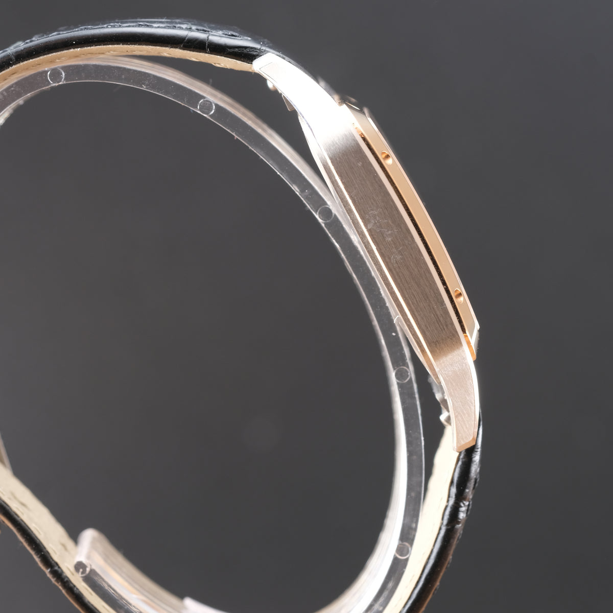 Close-up of a watch case showing a brushed rose gold metal finish and a black leather strap edge, photographed against a dark gray background.