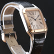 Cartier Santos watch with a silver-toned stainless steel case, gold-toned bezel, cream dial with Roman numerals, and a black leather strap, shown on a dark background.