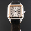 A Cartier Santos watch with a square stainless steel case, rose gold-toned bezel, white dial with black Roman numerals, and a black leather strap, shown against a dark gray background.